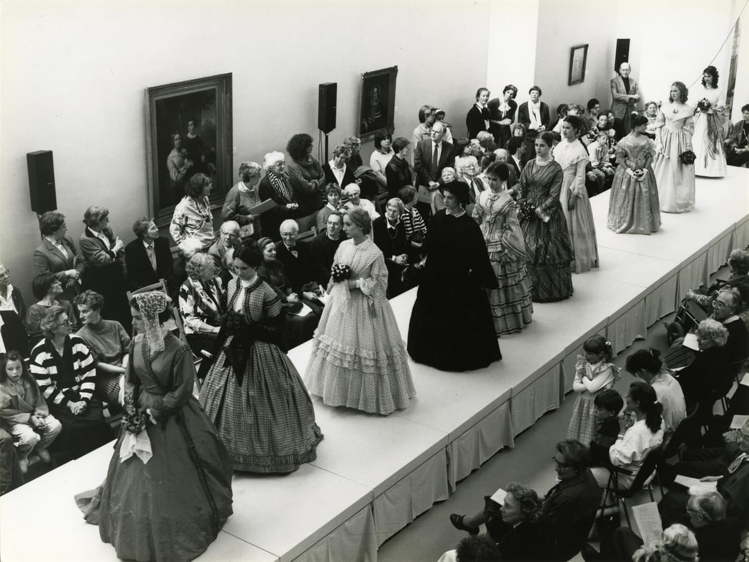 Zwart-witfoto van een modeshow in 1988 met de naam 'Ruisende Rokken' in het Centraal Museum, waarbij bezoekers kijken naar modellen gekleed in historische kostuums uit verschillende periodes die op de catwalk lopen.