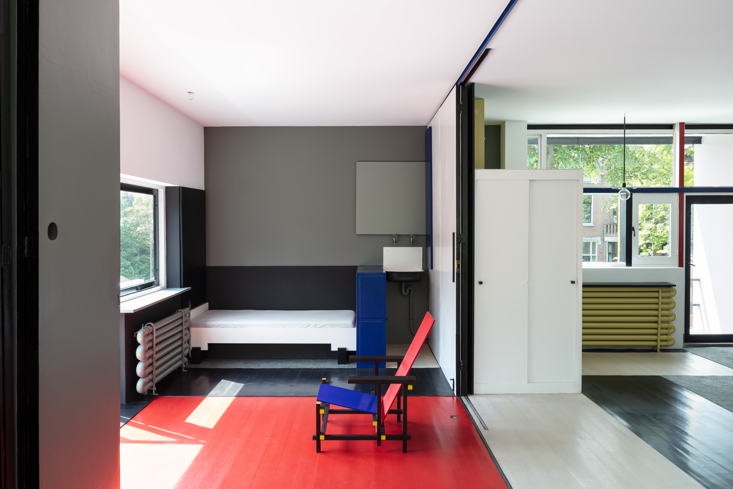 Modern interieur van het Rietveld Schröderhuis met een iconische rood-blauwe Rietveld stoel op een rode vloer, grijze en witte muren met zwart, geel en blauw accenten en een groot raam.
