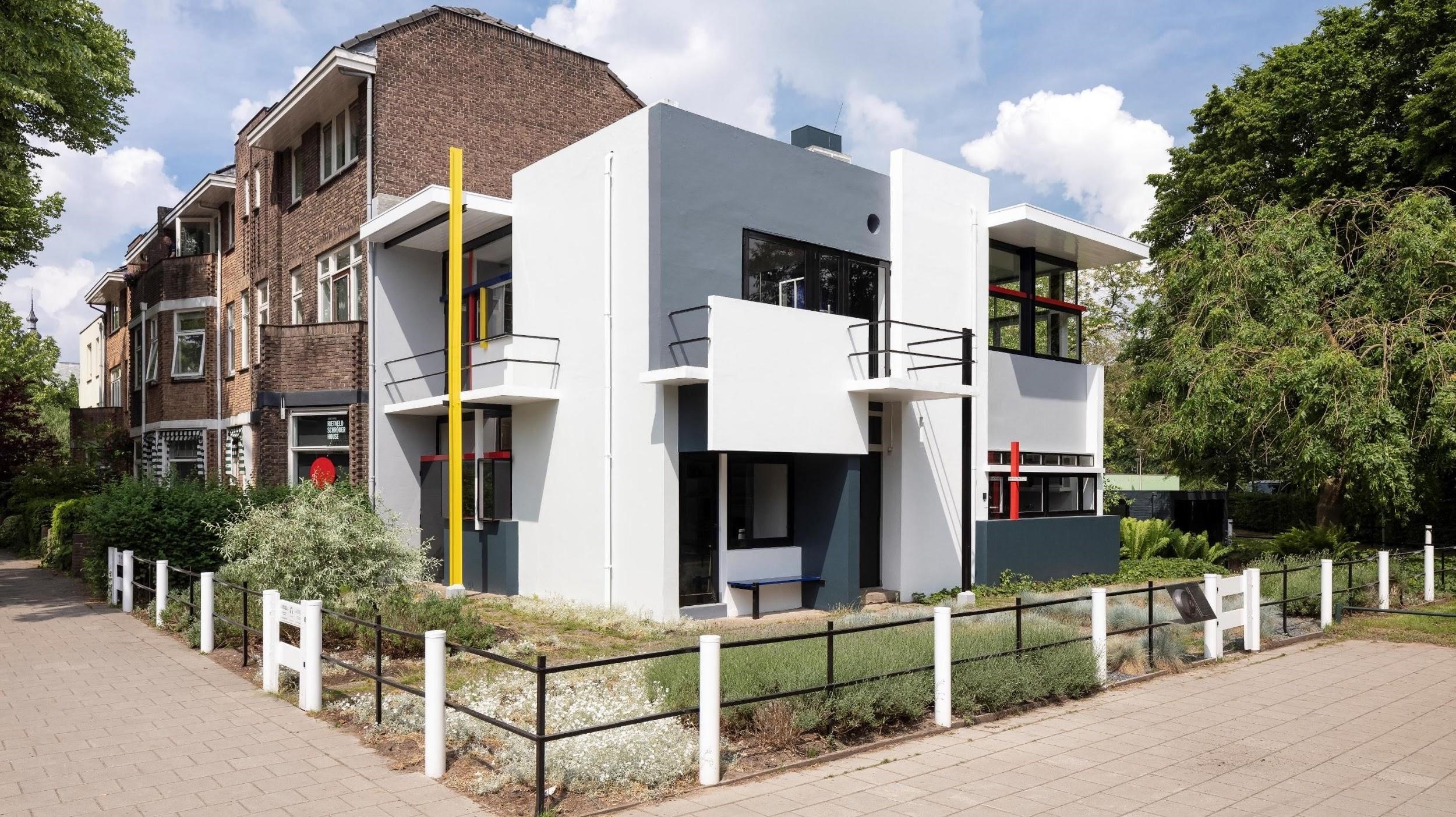 Foto van het Rietveld Schröderhuis: een modernistische witte villa met kenmerkende gele, rode en blauwe accenten van De Stijl, omgeven door groen en naast traditionele woningen.
