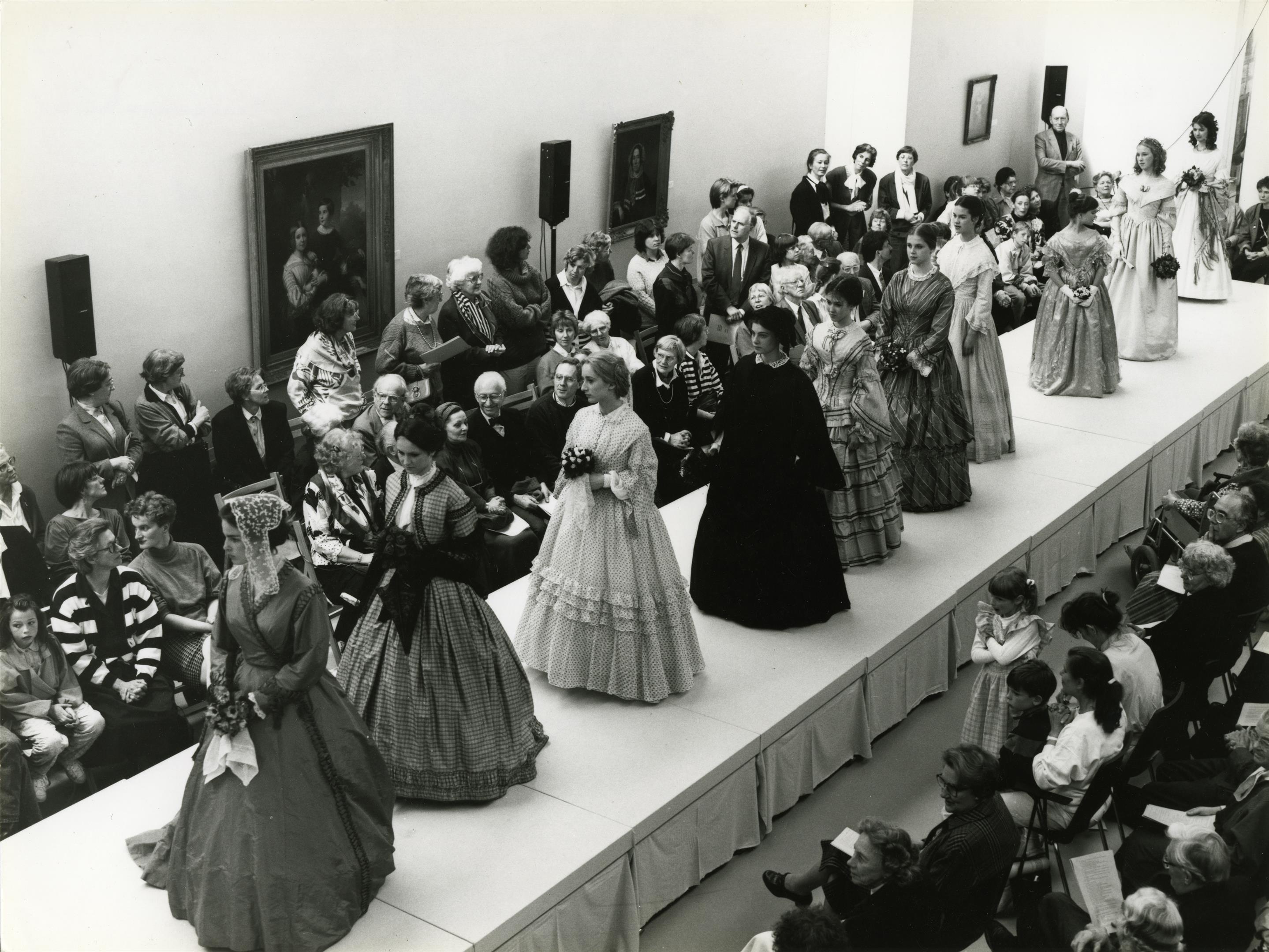 Zwart-witfoto van een modeshow in 1988 met de naam 'Ruisende Rokken' in het Centraal Museum, waarbij bezoekers kijken naar modellen gekleed in historische kostuums uit verschillende periodes die op de catwalk lopen.