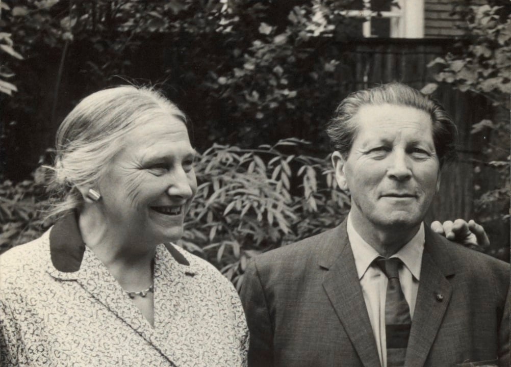 Zwart-witfoto van architect Gerrit Rietveld en ontwerpster Truus Schröder lachend in een tuin, gekleed in formele kleding uit het midden van de 20e eeuw.